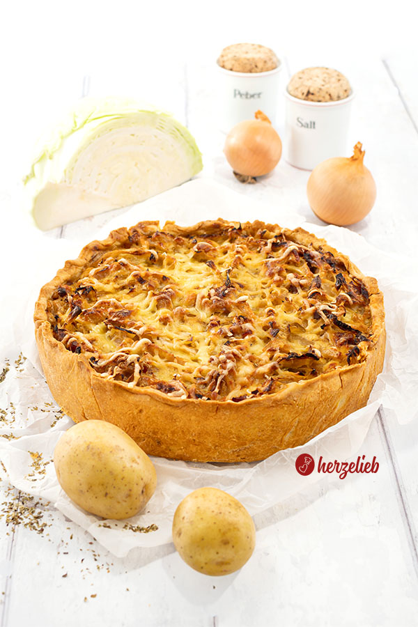 Ein Kohl-Kartoffel-Kuchen auf einem Stück Bakcpapier. Goldbraun und mit Käse überbacken. Im Vordergrund 2 Kartoffeln. Im Hintergrund ein Stück Kohl, 2 Zwiebeln sowie ein Pfefferstreuer und ein Salzstreuer.