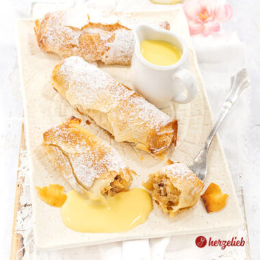 3 goldbraun gebackene Kuchenrollen nach einem Mini-Apfelstrudel Rezept von herzelieb liegen auf einem cremefarbenen Teller. Sie sind bestäubt mit Puderzucker, zwischen den kleine Kuchen steht eine kleine Kanne mit Vanillesoße und eine Zitronenhälfte liegt im Hintergrund.