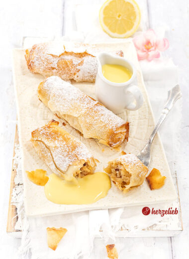 3 goldbraun gebackene Kuchenrollen nach einem Mini-Apfelstrudel Rezept von herzelieb liegen auf einem cremefarbenen Teller. Sie sind bestäubt mit Puderzucker, zwischen den kleine Kuchen steht eine kleine Kanne mit Vanillesoße und eine Zitronenhälfte liegt im Hintergrund.