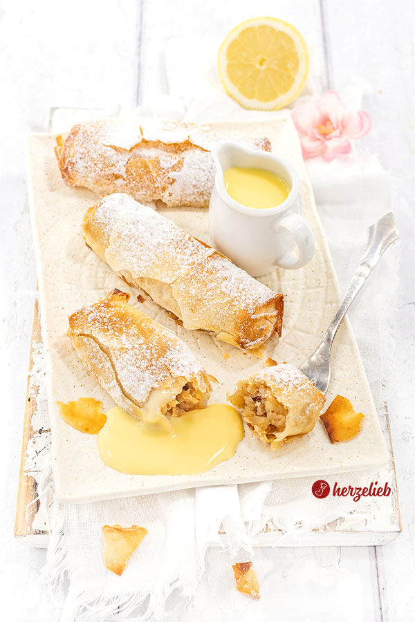 3 goldbraun gebackene Kuchenrollen nach einem Mini-Apfelstrudel Rezept von herzelieb liegen auf einem cremefarbenen Teller. Sie sind bestäubt mit Puderzucker, zwischen den kleine Kuchen steht eine kleine Kanne mit Vanillesoße und eine Zitronenhälfte liegt im Hintergrund.