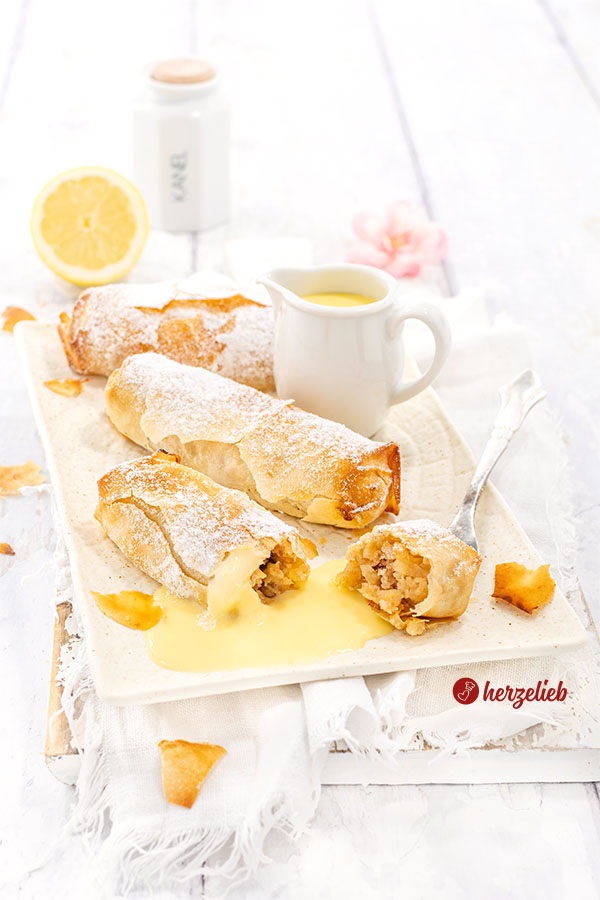 Drei kleine Apfelstrudel, bestäubt mit Puderzucker, liegen auf einem weißen rechteckigen Teller. Einer der Strudel ist angeschnitten, sodass die Apfelfüllung sichtbar wird, die von Vanillesoße umgeben ist. Im Hintergrund steht ein kleines Kännchen mit Vanillesoße und eine halbe Zitrone.