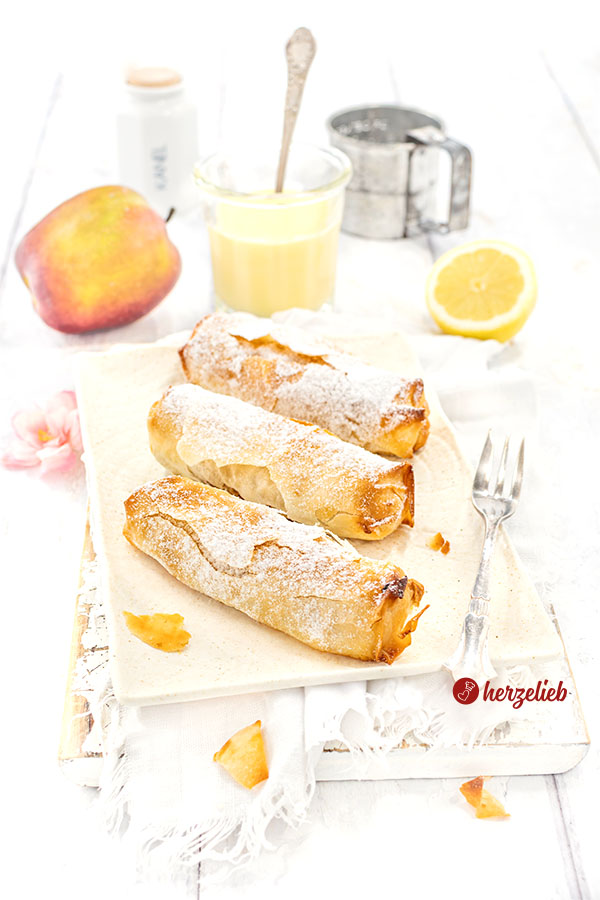 Drei frisch gebackene Mini-Apfelstrudel. Mit Puderzucker bestäubt, serviert auf einem einer cremefarbenen Porzellanplatte mit einer knitterigen Tuch, dazu ein Glas Vanillesauce und frische Zutaten wie Apfel und Zitrone im Hintergrund.