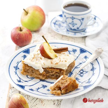 Ein Stück dänischer Apfelkuchen nach einem Svampet Æblekage Rezept aus Dänemark von herzelieb. Der Blechkuchen hat ein Frischkäse-Topping und ist dekoriert mit einem Apfel und einer Zimtstange. Ein Stück von dem Kuchenstück ist auf einer Gabel.