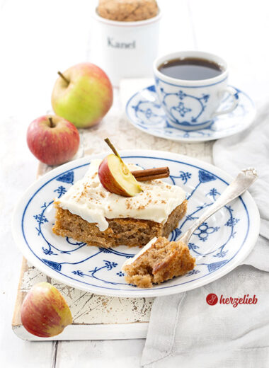Ein Stück dänischer Apfelkuchen nach einem Svampet Æblekage Rezept aus Dänemark von herzelieb. Der Blechkuchen hat ein Frischkäse-Topping und ist dekoriert mit einem Apfel und einer Zimtstange. Ein Stück von dem Kuchenstück ist auf einer Gabel.