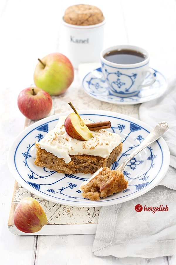 Ein Stück dänischer apfelkuchen nach einem Svampet Æblekage Rezept aus Dänemark von herzelieb. Der Blechkuchen hat ein Frischkäse-Topping und ist dekoriert mit enem Apfel und einer Zimstange. Ein Stück von dem Kuchenstück ist auf einer Gabel.