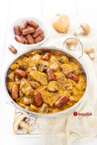 Eine Edelstahl-Servierschale mit einem Wikingertopf aus Dänemark nach einem Vikingegryde Rezept von herzelieb. Im Hintergrund eine Schale mit Cocktailwürstchen, Zwiebeln und braune Champignons.