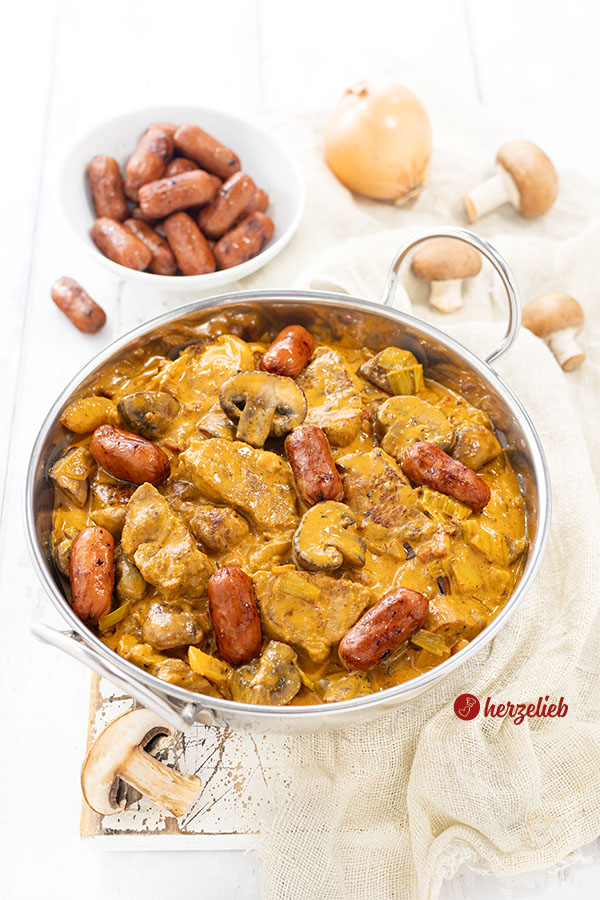 Eine Edelstahl-Servierschale mit einem Wikingertopf aus Dänemark nach einem Vikingegryde Rezept von herzelieb. Im Hintergrund eine Schale mit Cocktailwürstchen, Zwiebeln und braune Champignons.