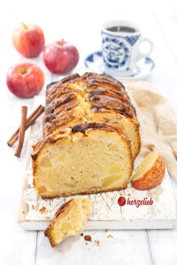 Angeschnittener Apfelkuchen aus der Kastenform. Dekoriert mit Äpfeln und Zimtstangen. Im Hintergrund eine Tasse Kaffee.