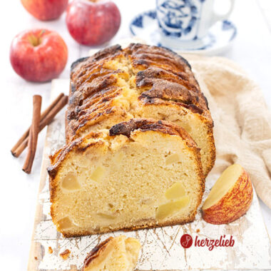 Angeschnittener Apfelkuchen aus der Kastenform. Dekoriert mit Äpfeln und Zimtstangen. Im Hintergrund eine Tasse Kaffee.