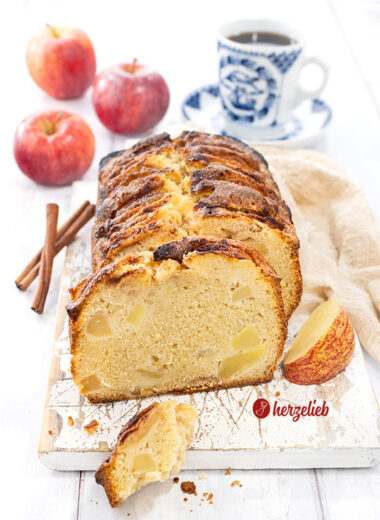 Angeschnittener Apfelkuchen aus der Kastenform. Dekoriert mit Äpfeln und Zimtstangen. Im Hintergrund eine Tasse Kaffee.