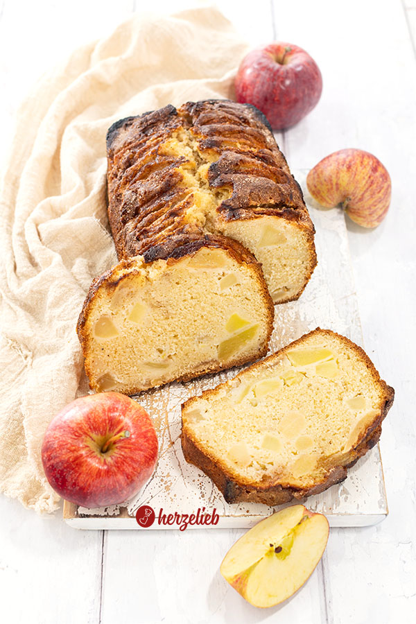 Knuspriger, angeschnittener Apfelkuchen aus der Kastenform auf einem weißen Holzbrett. Rundherum liegen Äpfel.