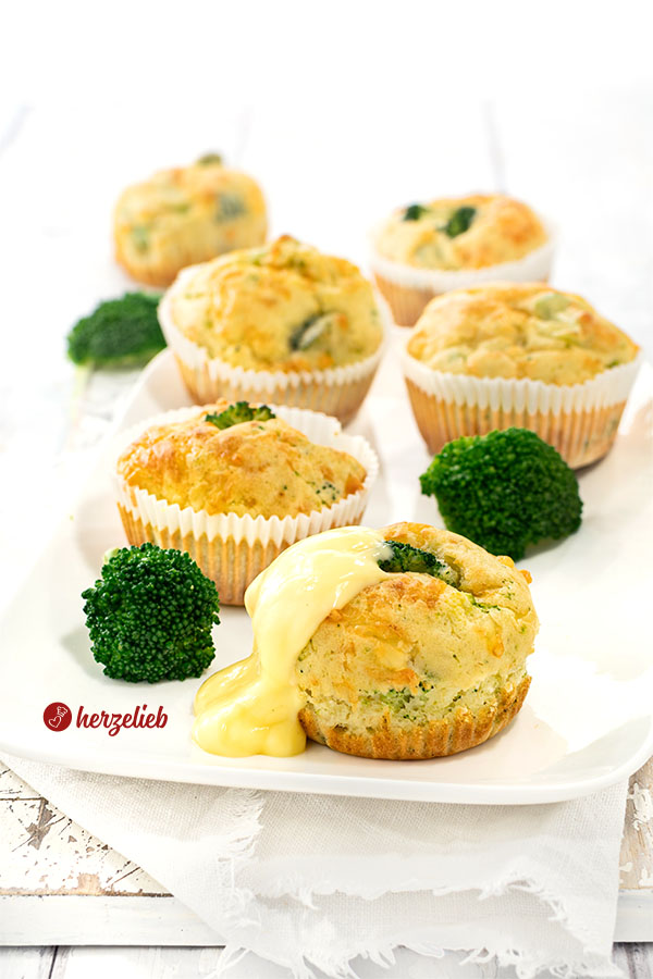 Goldbraun gebackene, herzhafte Muffins nach einem Brokkoli-Käse-Muffins Rezept von herzelieb. Im Vordergrund einer der Brokkolimuffins mit Sauce Hollandaise. Zwischen den Muffins liegen Brokkoli-Röschen.