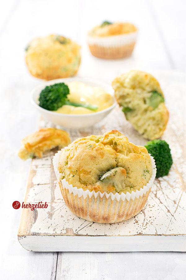 3 Brokkolimuffins nach einem Brokkoli-Käse-Muffin Rezept mit Käse auf einem Holzbrett. Hinter dem vorderen der Käsemuffins mit Brokkoli steht eine Schale mit Sauce Hollandaise, an dem ein halber Käsemuffin lehnt.