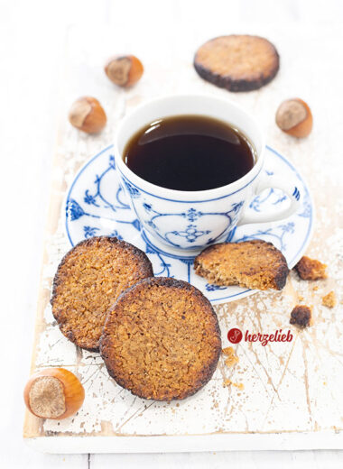 2 ganze Nussplätzchen nach einem Hasselnøddesmåkager Rezept aus Dänemark vor einer blauweißen Tasse. Auf der Untertasse liegt ein angebissenes Plätzchen. Die Haselnusskekse haben einen dunklen Rand. Im Hintergrund ein weiterer Keks. Dekoriert mit ganzen Haselnüssen.