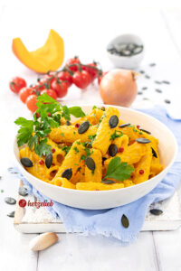 Kürbis-Feta-Pasta in einer weißen Schale. Das Nudelgericht ist durch die Kürbissoße mit Fetakäse intensiv gelb bis Orange und ist angerichtet mit rosa Pfefferbeeren, Kürbiskernen und Petersilie. Im Hintergrund Tomaten, eine Zwiebel, ein Stück Kürbis und eine Schale Kürbiskerne.