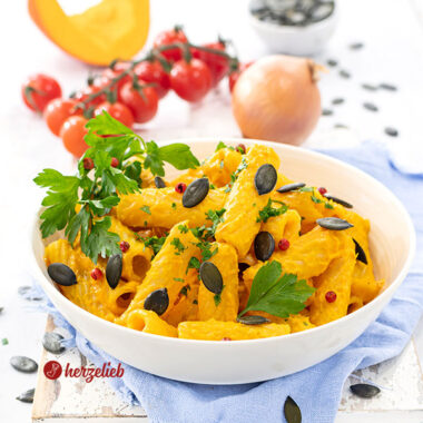 Kürbis-Feta-Pasta in einer weißen Schale. Das Nudelgericht ist durch die Kürbissoße mit Fetakäse intensiv gelb bis Orange und ist angerichtet mit rosa Pfefferbeeren, Kürbiskernen und Petersilie. Im Hintergrund Tomaten, eine Zwiebel, ein Stück Kürbis und eine Schale Kürbiskerne.