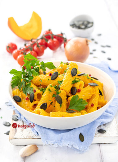 Kürbis-Feta-Pasta in einer weißen Schale. Das Nudelgericht ist durch die Kürbissoße mit Fetakäse intensiv gelb bis Orange und ist angerichtet mit rosa Pfefferbeeren, Kürbiskernen und Petersilie. Im Hintergrund Tomaten, eine Zwiebel, ein Stück Kürbis und eine Schale Kürbiskerne.