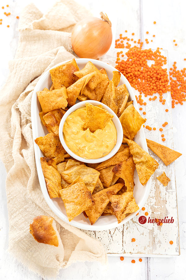 Eine ovale Porzellanplatte mit knusprigen Chips nach einem Linsenchips Rezept von herzelieb.  In der Mitte der Platte steht Schälchen mit einem Dip. Im Hintergrund liegen verstreut rote Linsen, daneben eine Zwiebel. Links ein beigefrbenes Tuch.