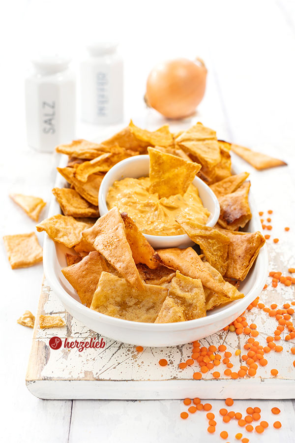 Eine flache ovale Porzellanschale mit Chips nach einem Linsenchips Rezept von herzelieb. In der Mitte ein Schälchen mit einem orangefarbenen Dip. im Hintergrund ein Salzstreuer, ein Pfefferstreuer und eine Zwiebel.