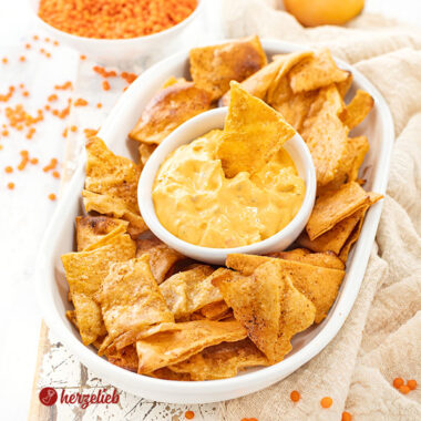 Eine flache, ovale Porzellanplatte mit Linsenchips, in der Mitte ein weiße Schale mit einem orangfarbenen Dips. Im Hintergrund eine Zwiebel und eine Schale mit roten Linsen.