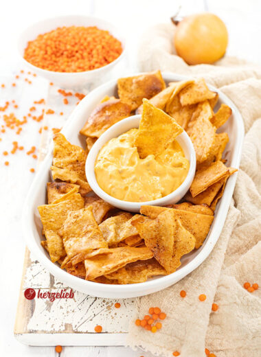 Eine flache, ovale Porzellanplatte mit Linsenchips, in der Mitte ein weiße Schale mit einem orangfarbenen Dips. Im Hintergrund eine Zwiebel und eine Schale mit roten Linsen.