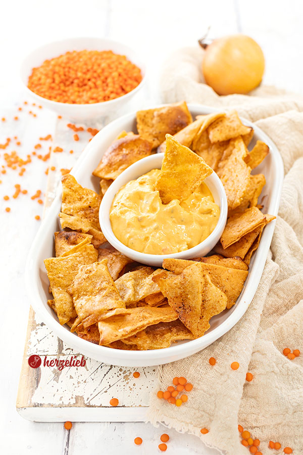 Eine flache, ovale Porzellanplatte mit Linsenchips, in der Mitte ein weiße Schale mit einem orangfarbenen Dips. Im Hintergrund eine Zwiebel und eine Schale mit roten Linsen.