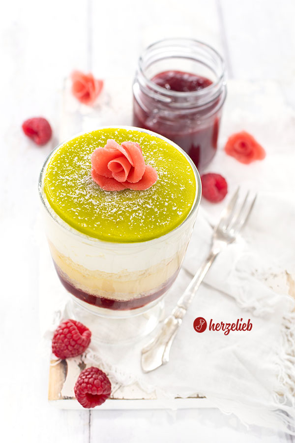 Ein buntes Schichtdesserts nach einem Prinsesstårta Dessert im Glas von herzelieb mit 5 Schichten, einem grünen Marzipandeckel, der mit Puderzucker bestäubt und einer rosa Rose dekoriert ist. Im Hintergrund ein Glas Himbeermarmelade. Die schwedische Torte als Nachtisch.