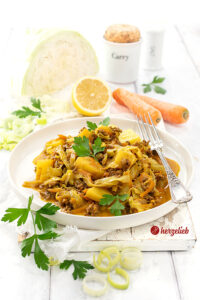 Ein weißer Telller mit Indianergryde aus Dänemark. Zu sehen sind im Gericht Kartoffeln,  Weißkohlstreifen und geraspelte Karotten. Dekoriert mit Petersilie. Auf dem Tellerrand eine Gabel. Im Hintergrund ein Weißkohl, Zitrone, Möhren und 2 Gewürzgefäße aus Porzellan. 