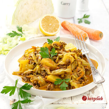 Ein weißer Telller mit Indianergryde aus Dänemark. Zu sehen sind im Gericht Kartoffeln, Weißkohlstreifen und geraspelte Karotten. Dekoriert mit Petersilie. Auf dem Tellerrand eine Gabel. Im Hintergrund ein Weißkohl, Zitrone, Möhren und 2 Gewürzgefäße aus Porzellan.