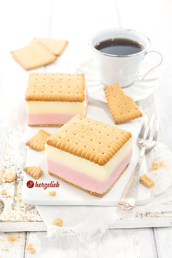 Zwei Stücke vom Joghurt-Wackel-Kuchen auf einem weißen Brett mit einer Tasse Kaffee und einer Kuchengabel. Die Kuchenstücke habe eine weiße und eine rosafarbene Schicht und einen Boden und eine Deckel aus Butterkeks.