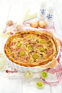 Eine goldbraun gebackene Käse-Lauch-Tarte mit Hackfleisch. Angerichtet in einer weißen Tarteform, steht auf einem weißen Holzbrett, auf dem ein weiß-rotes Geschirrtuch liegt. Im Hintergrund Lauch, Zwiebeln und je ein Salz- und Pfefferstreuer.