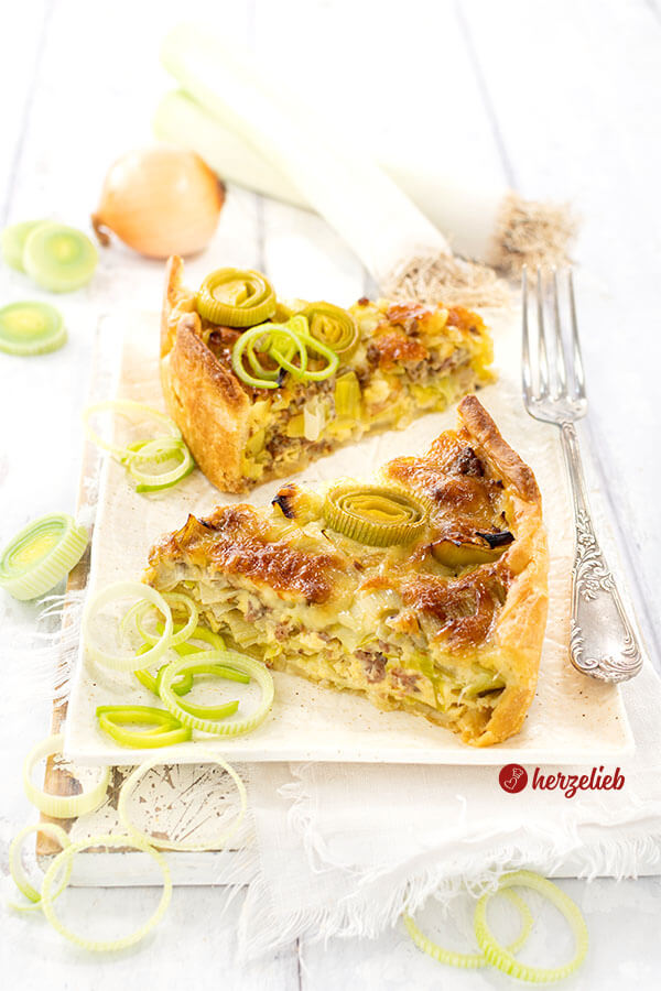 Zwei Stücke von einer goldbraun gebackenen Käse-Lauch-Tarte mit Hackfleisch. Angerichtet mit frischen Lauchringen. Auf dem Tellerrand eine Gabel und im Hintergrund zwei Lauchstangen und eine Zwiebel.