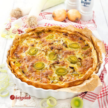 Eine goldbraun gebackene Käse-Lauch-Tarte mit Hackfleisch. Angerichtet in einer weißen Tarteform, steht auf einem weißen Holzbrett, auf dem ein weiß-rotes Geschirrtuch liegt. Im Hintergrund Lauch, Zwiebeln und je ein Salz- und Pfefferstreuer.