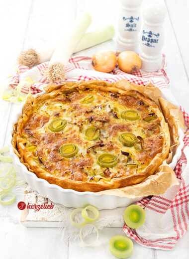 Eine goldbraun gebackene Käse-Lauch-Tarte mit Hackfleisch. Angerichtet in einer weißen Tarteform, steht auf einem weißen Holzbrett, auf dem ein weiß-rotes Geschirrtuch liegt. Im Hintergrund Lauch, Zwiebeln und je ein Salz- und Pfefferstreuer.