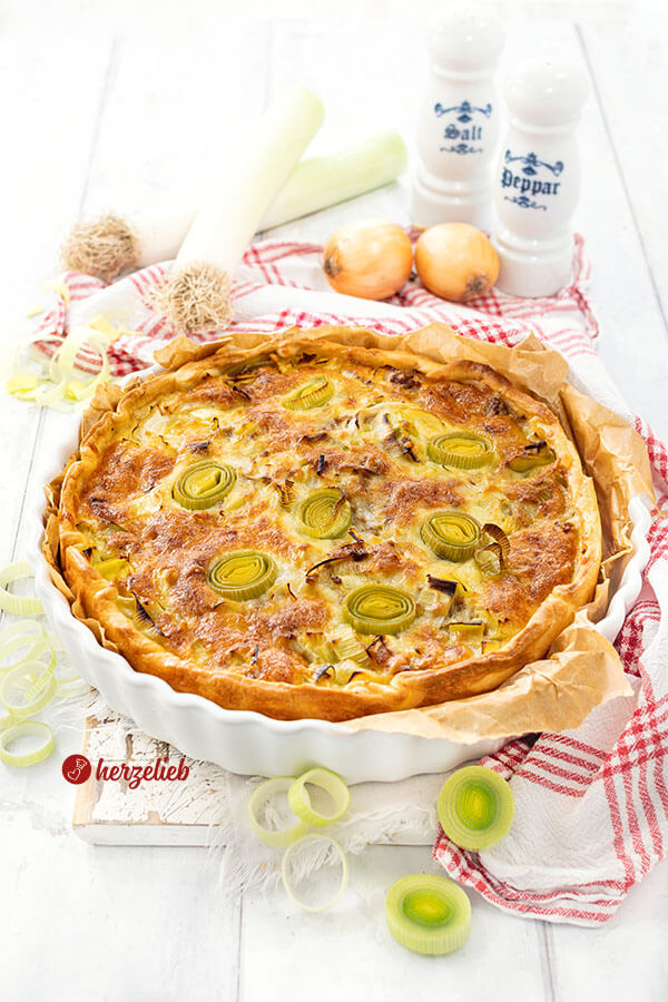 Eine goldbraun gebackene Käse-Lauch-Tarte mit Hackfleisch. Angerichtet in einer weißen Tarteform, steht auf einem weißen Holzbrett, auf dem ein weiß-rotes Geschirrtuch liegt. Im Hintergrund Lauch, Zwiebeln und je ein Salz- und Pfefferstreuer.