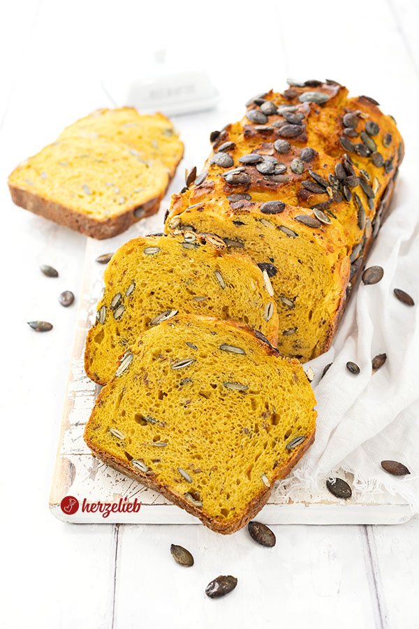 Ein angeschnittenes Kürbisbrot nach einem Kürbis-Buttermilchbrot Rezept von herzelieb.Das Brot ist goldbraun gebacken und mit Kürbiskernen bestreut. Vor dem Brotlaib liegen zwei Scheiben davon. Im Hintergrund zwei weitere Scheiben.