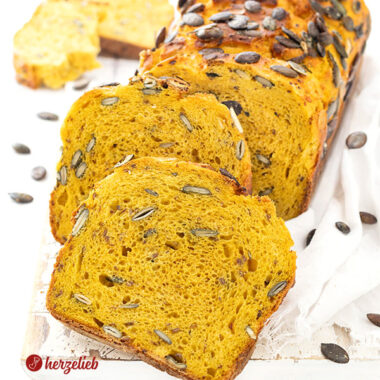 Ein saftiges und angeschnittenes Kürbisbrot oder Buttermilchbrot nach einem Kürbis-Buttermilchbrot Rezept von herzelieb. Zwei Scheiben davon lieben vor dem angeschnittenen Brotlaib.