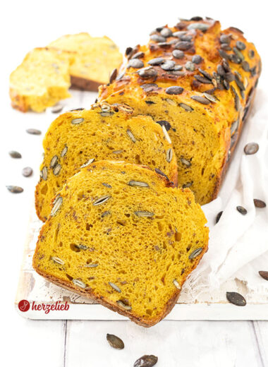 Ein saftiges und angeschnittenes Kürbisbrot oder Buttermilchbrot nach einem Kürbis-Buttermilchbrot Rezept von herzelieb. Zwei Scheiben davon lieben vor dem angeschnittenen Brotlaib.
