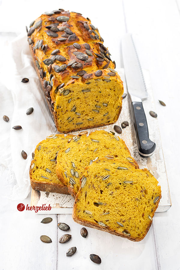 Ein angeschnittener Laib Kürbis-Buttermilchbrot mit drei Scheiben, die davor liegen. Das Brot ist bestreut mit Kürbiskernen. Rechts im Hintergrund ein Brotmesser. 