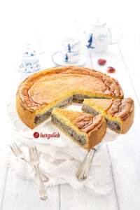 Angeschnittener Mohnkuchen auf einem Cakestand. Der Kuchen hat drei Schichten - Mürbeteigboden, Mohnschicht mit Grieß und Puddingguss. Im Hintergrund blauweßes