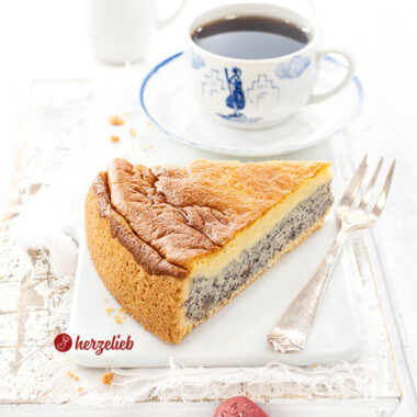 Ein Stück Kuchen nach einem Mohnkuchen Rezept von herzelieb auf einem weißen Brett. Neben dem Kuchenstück eine Gabel, im Hintergrund eine Tasse Kaffee.