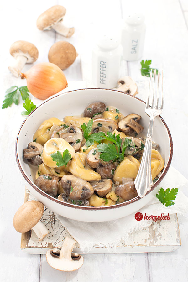 Eine Schüssel mit einem Nudelgericht nach einem Pilz-Tortellini-Rezept von herzelieb. Dekoriert mit Petersilie. Auf dem Schüsselrand eine Gabel. Im Hintergrund eine Zwiebel, Champignons und ein Salzstreuer und ein Pfefferstreuer.