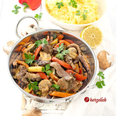Pfannengericht nach einem Steakpfanne Rezept von herzelieb in einer silberfarbenen Servierpfanne. Im Gericht sieht man Paprika, Zucchni, Teakstreifen und Pilze iner cremigen Senfsoße. Im Hintergrund eine Schüssel mit Kartoffelbrei.