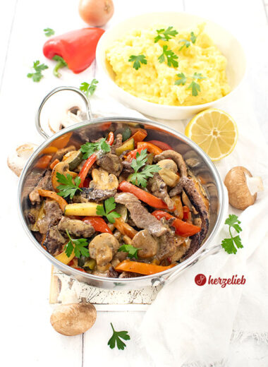 Pfannengericht nach einem Steakpfanne Rezept von herzelieb in einer silberfarbenen Servierpfanne. Im Gericht sieht man Paprika, Zucchni, Teakstreifen und Pilze iner cremigen Senfsoße. Im Hintergrund eine Schüssel mit Kartoffelbrei.