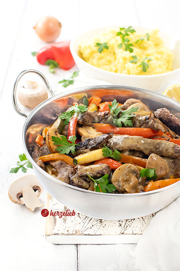 Eine Servierpfanne mit einer Steakpfanne in cremiger Senfsoße. Im Hintergrund Kartoffelbrei und ein Paprika