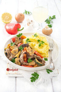 Ein Teller mit Steakpfanne mit Senfsoße zu Kartoffelbrei. Im Hintergrund Pilze, Paprika, Petersilie, eine Zwiebel und eine Zitrone. Auf dem Tellerrand eine Gabel.