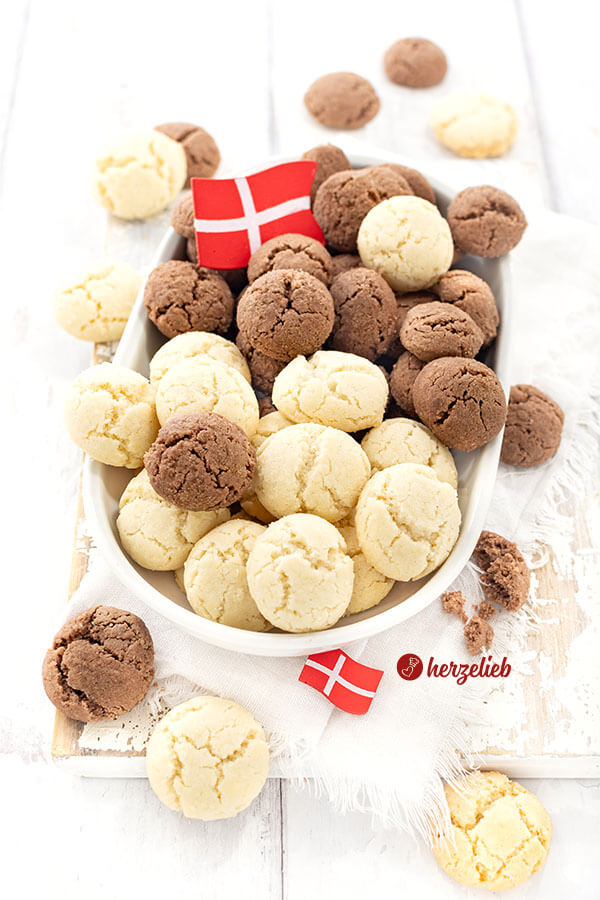 Helle und dunkle Plätzchen aus Dänemark nach einem Fedtkager Rezept von herzelieb auf einer weißen, ovalen Porzellanplatte. In den Plätzchen steckt ein Dannebrog, vor der Platte liegt auch einer und ringsherum sind weitere Kekse verteilt.