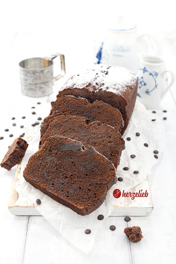 Ei aufgeschnittener, mit Puderzucker bestäubter Schokokuchen nach einem Frischkäse-Schokoladenkuchen Rezept von herzelieb auf einem weißen Brett. Vor dem Kuchen liegen versetzt 3 Scheiben davon.