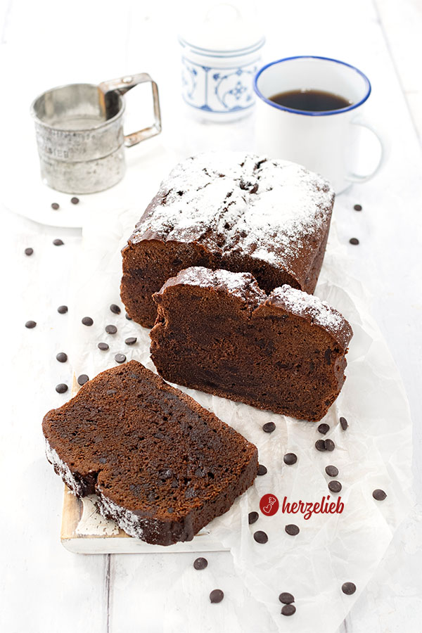 Aufgeschnittener, saftiger Schokokuchen mit Schokoladenstücken nach einem Frischkäse-Schokoladenkuchen Rezept von herzelieb. Der Kuchen ist mit Puderzucker bestäubt. Eine Scheibe teht aufrecht davor, die andere andere liegt links davor.