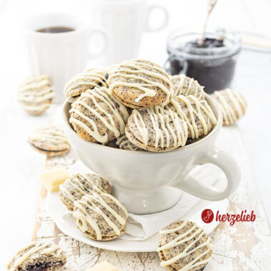 Eine große Tasse, gefüllt mit gefüllten Mohnplätzchen nach einem Germknödel Plätzchen Rezept von herzelieb. Die Plätzchen sind mit weißer Kuvertüre verziert. . Im Hintergrund ein Glas mit Pflaumenmus und zwei Becher Kaffee.